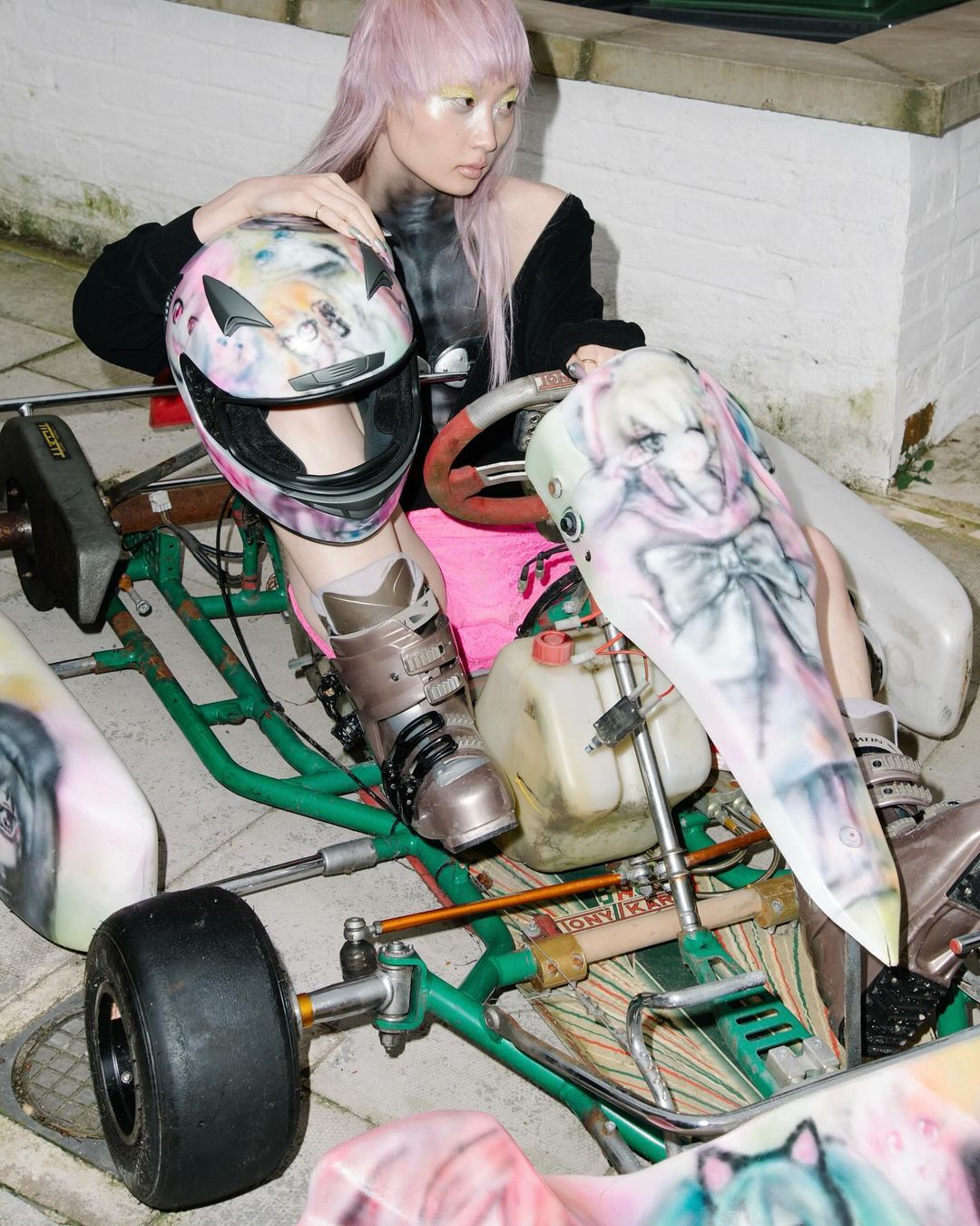 Asian woman with pink hair in sporty outfit poses with a customized go-kart and helmet, showcasing unique style and automotive culture.