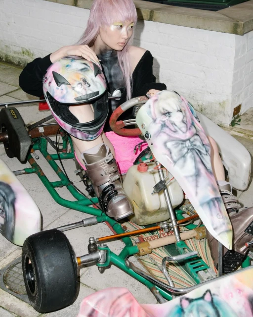 Asian woman with pink hair in sporty outfit poses with a customized go-kart and helmet, showcasing unique style and automotive culture.