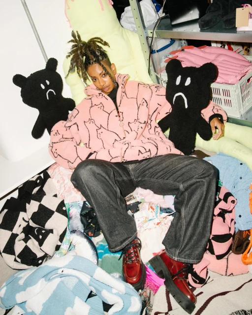 Young adult woman in a pink cat-print hoodie sitting among textile piles and holding plush bear toys, with a shelf unit in background.
