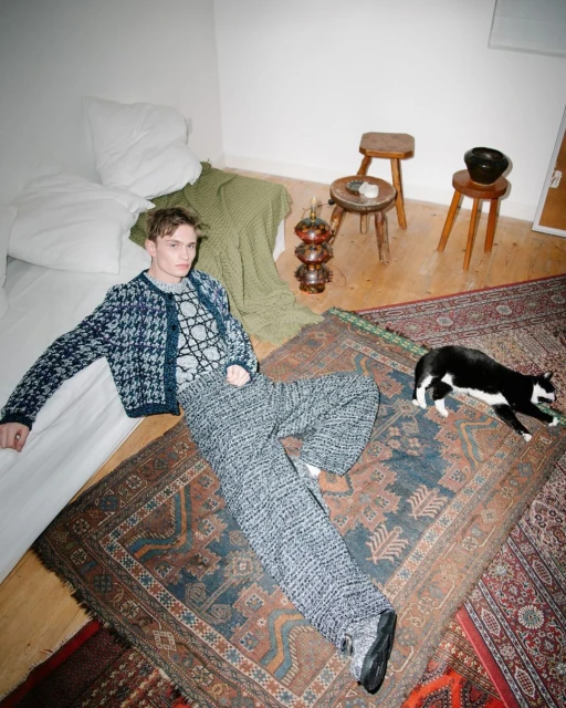 Young adult poses on an antique rug in a bedroom with a black-and-white cat, pillows, and wooden stools. Soft, diffused natural light.