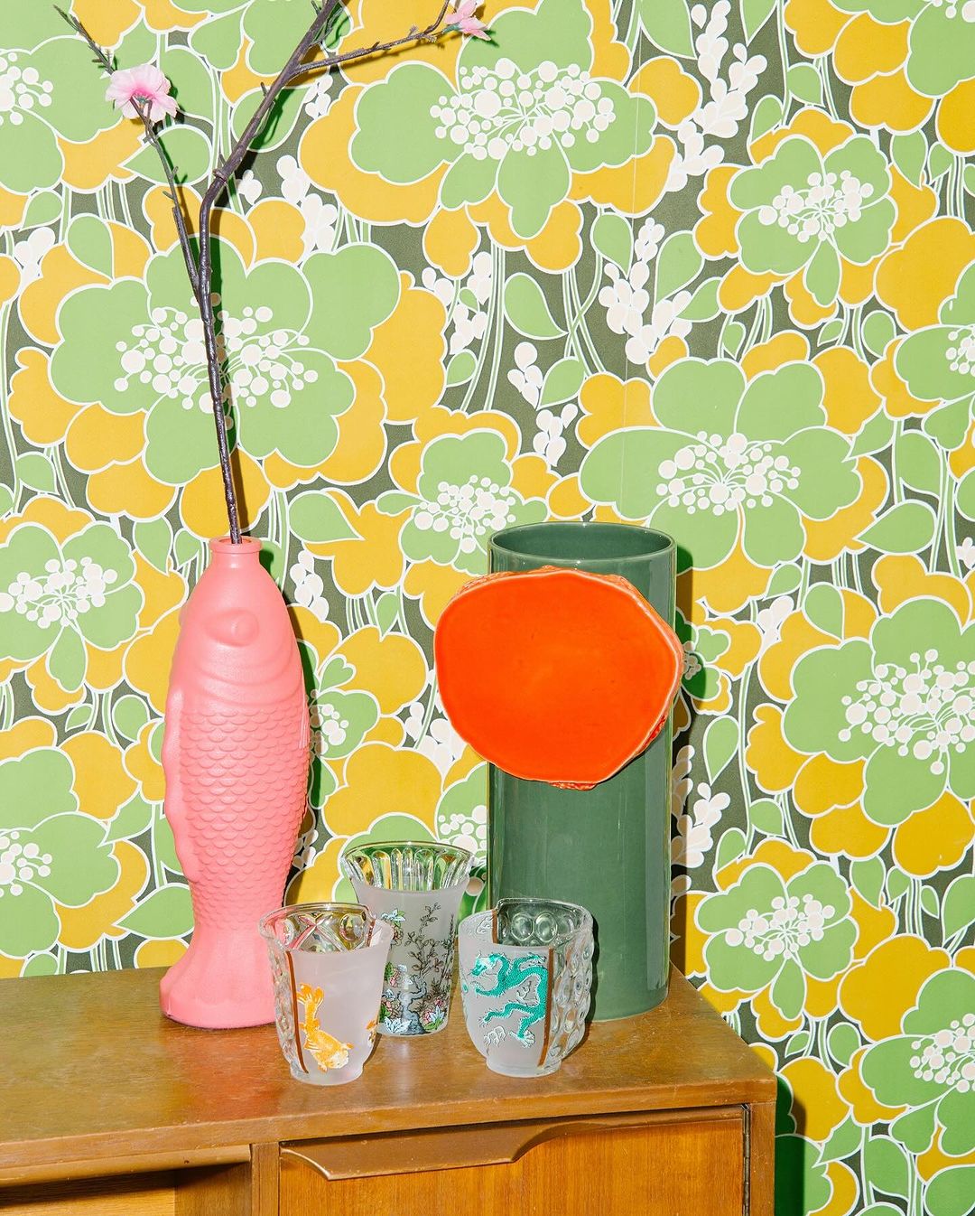 Still life with floral wallpaper, a pink fish vase holding floral branches, glassware, and a green vase with an orange disc.