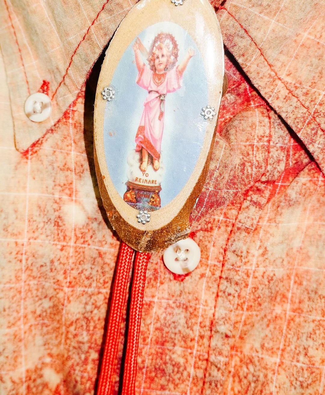 Vintage photo of an ornate bolo tie on a light orange button-down shirt with red stitching, featuring a childhood depiction of a person.