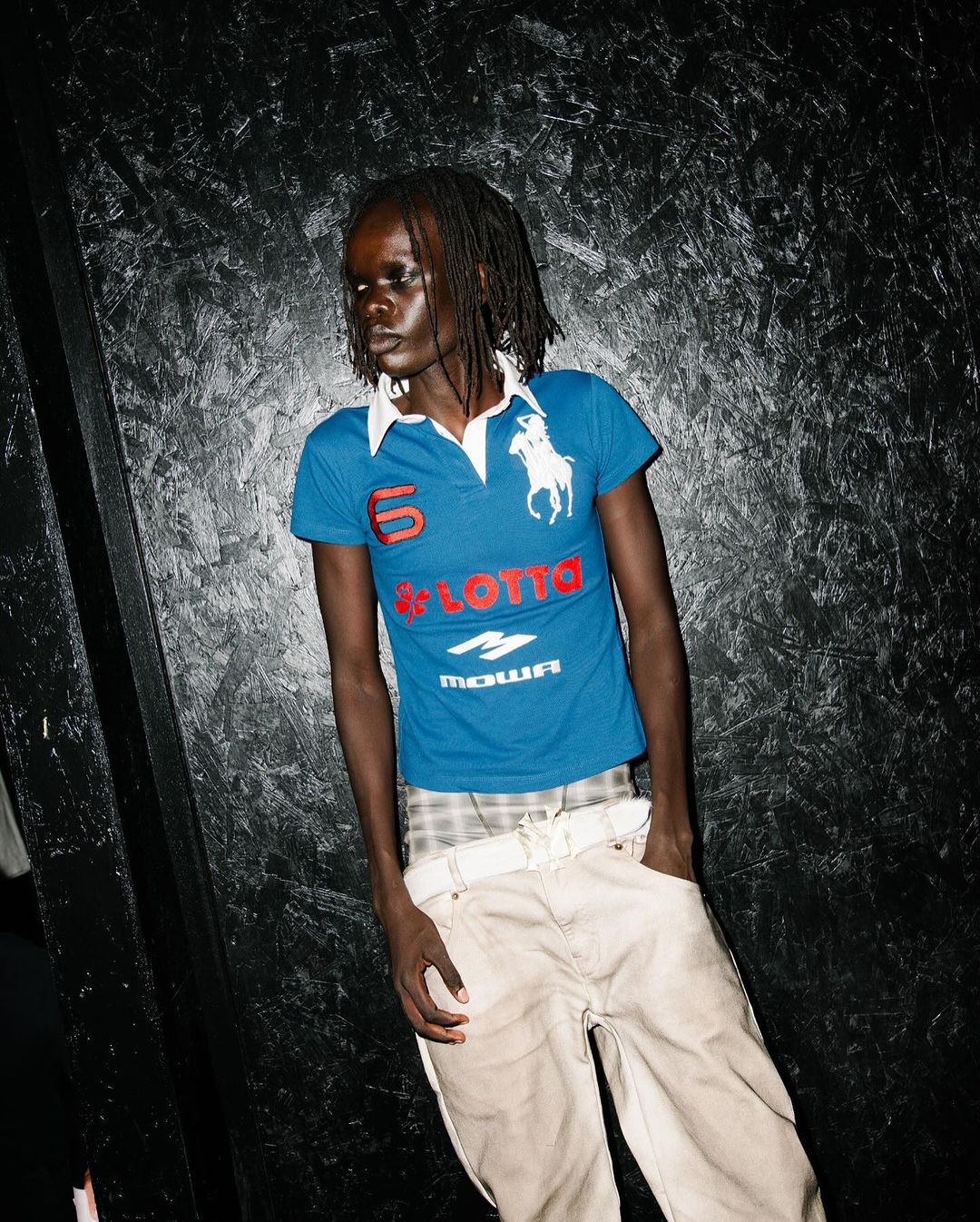 A dark-skinned person with dreadlocks poses against a dark textured wall, wearing a blue graphic tee and light-colored pants with hands in pockets.