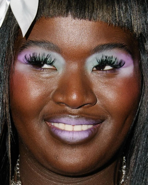 Close-up portrait of a Black woman with dramatic makeup, including lilac lipstick and pastel eyeshadow. Hair is dark with a white ribbon.