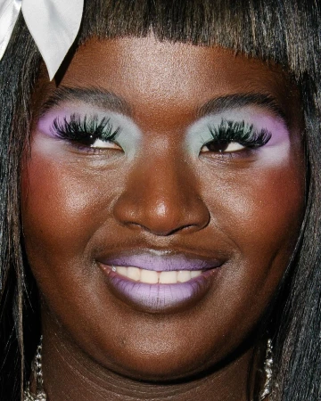 Close-up portrait of a Black woman with dramatic makeup, including lilac lipstick and pastel eyeshadow. Hair is dark with a white ribbon.