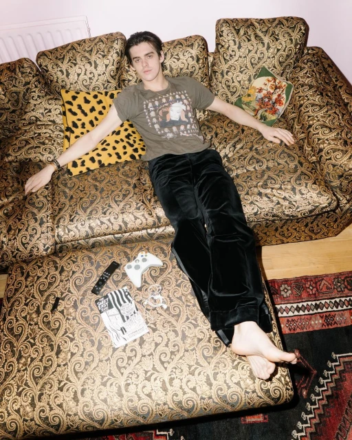 Young man lounging on a patterned couch in olive t-shirt and black velvet pants, near game controller and magazine. Interior shot.