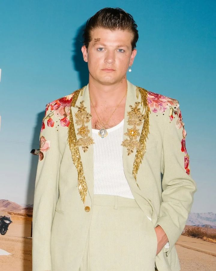 Male model in pale green suit with floral embroidery, posing against a blue sky and desert background, showcasing fashion and style.