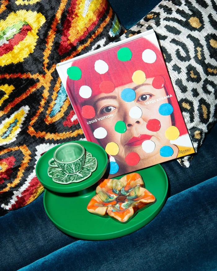 An art book and green decorative objects resting on a textured blue velvet sofa with patterned cushions.
