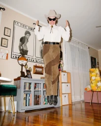 Man wearing a cowboy hat and sunglasses jumping in a living room photographed by Bob Foster.