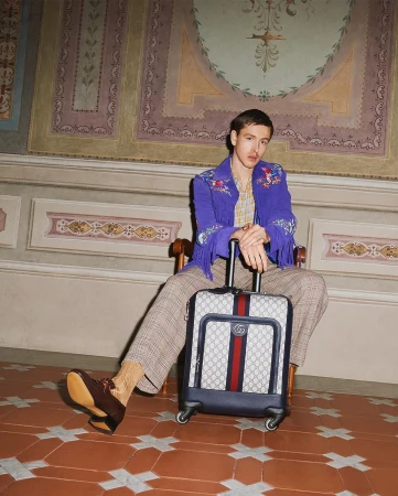Man wearing a purple fringed jacket seated in a chair holding a patterned suitcase with red and blue stripes.