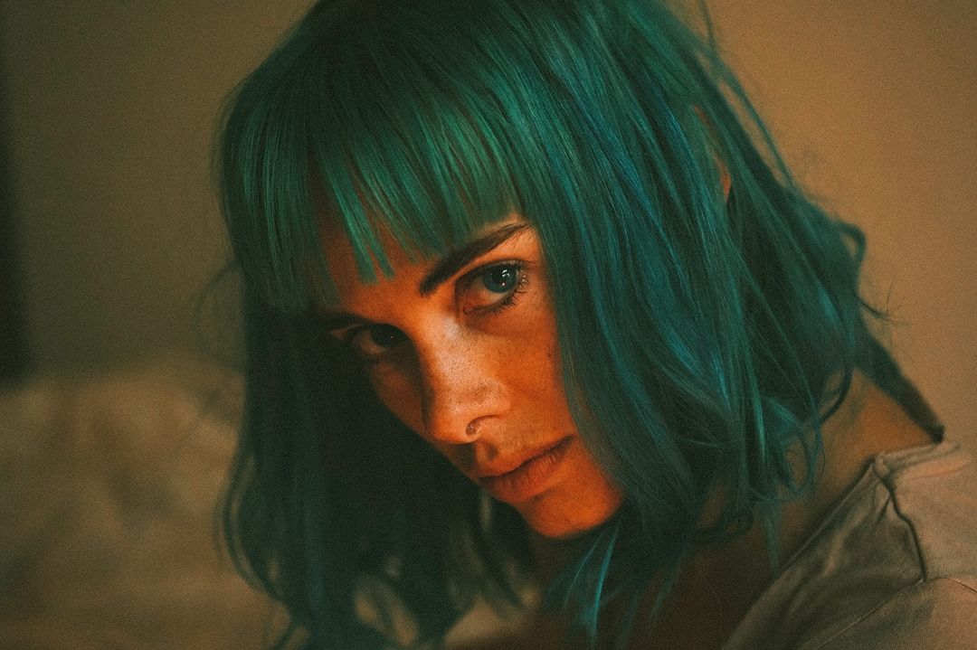Close-up portrait of a woman with teal hair, looking at the camera with a serious expression against a neutral, warm-toned background.