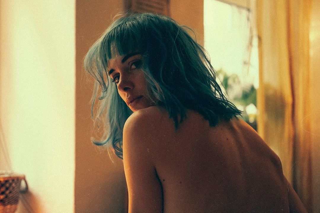 Woman with blue hair and bare back looks over her shoulder in a medium shot in a warm indoor setting, filtered with a vintage effect.