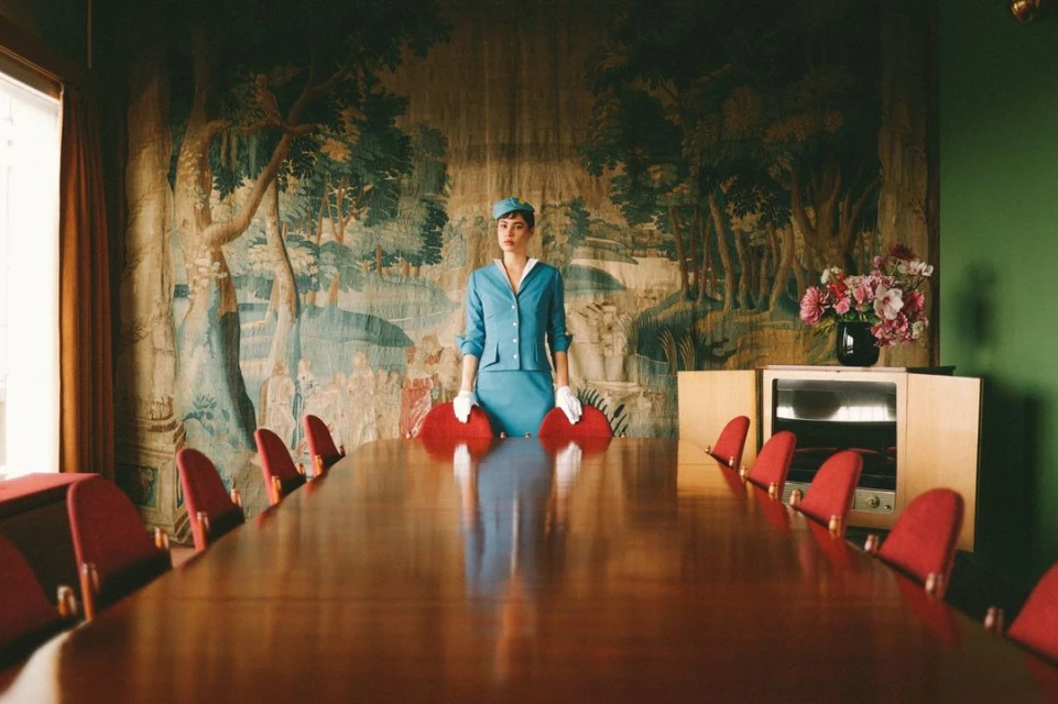 Elegant vintage-style shot of a woman in blue suit, hat, and gloves standing by a long conference table against a tapestry backdrop.