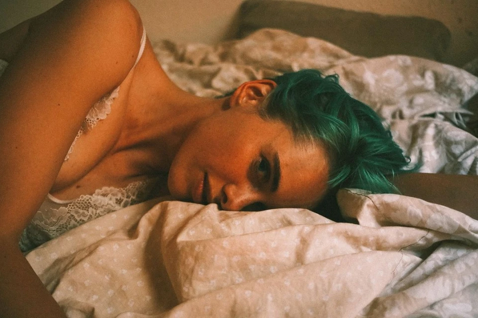 Close-up portrait of a woman with dyed green hair lying on a bed covered with a patterned duvet. Soft, warm lighting creates a cozy mood.