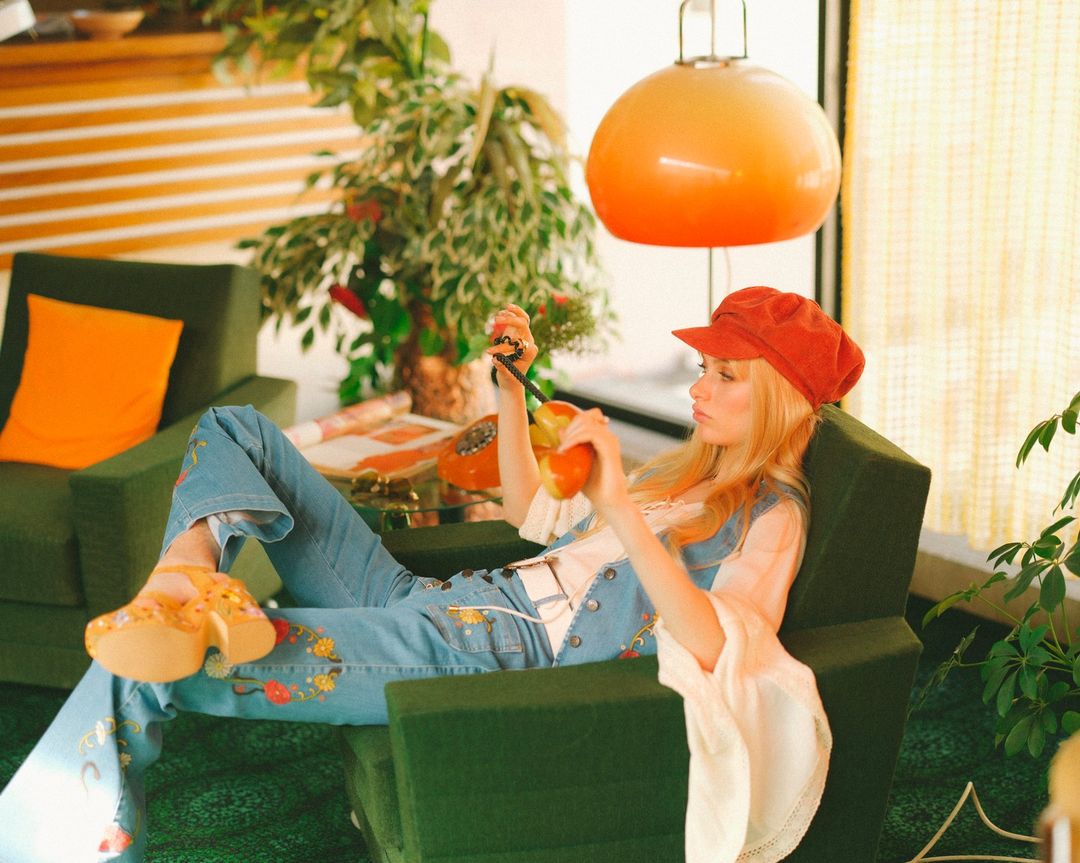 A woman in 1970s fashion reclining in a green chair, holding a rotary phone receiver, in a room with retro decor.