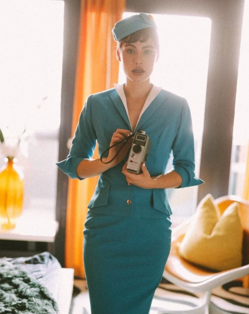 Elegant woman in a teal suit and matching hat stands indoors, holding a vintage camera, creating a retro and stylish portrait.