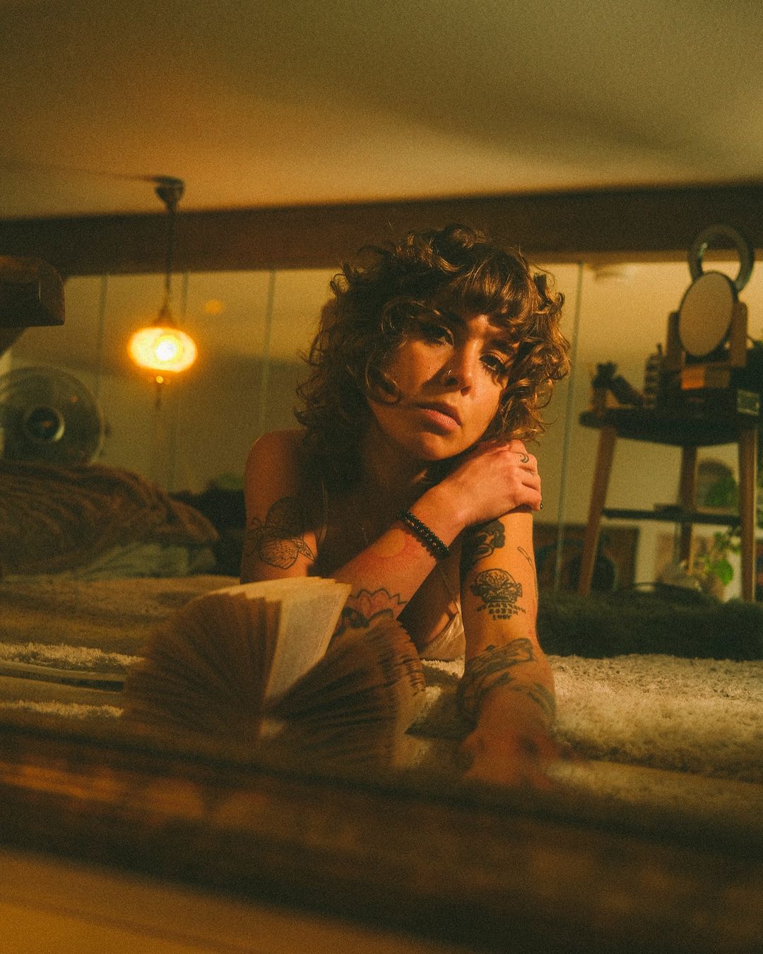Indoor shot of a curly-haired woman lying on a bed with tattoos, looking at an open book in a dimly lit room with warm tones.