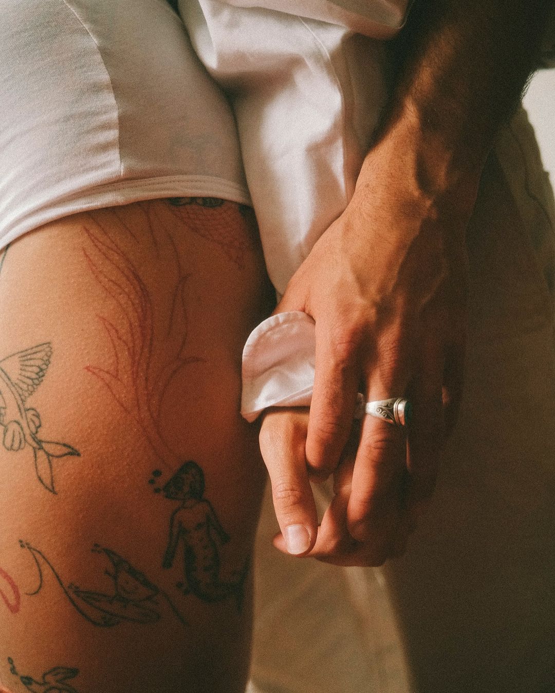 Close-up shot of a person with tattoos on their leg, holding hands with another person wearing a ring and a white shirt.