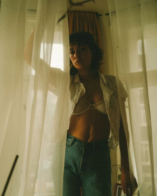 Young woman poses indoors in blue jeans, white shirt and bra behind semi-sheer curtain, with a warm color grade, and natural window lighting.