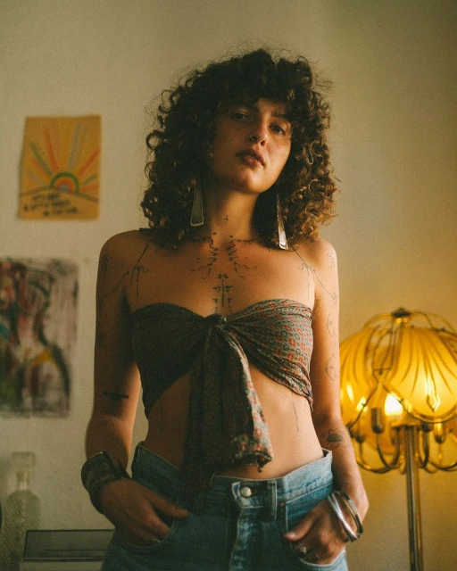 Indoor portrait of a woman with curly hair wearing a tied scarf top and jeans, standing in a warmly lit room with artwork and a lamp.