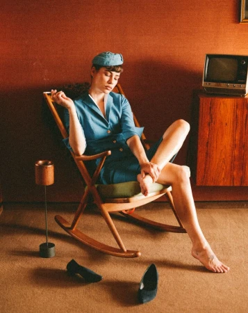 Woman in blue dress sitting in rocking chair, holding a cigarette, with vintage TV and pair of shoes, against a warm toned backdrop.
