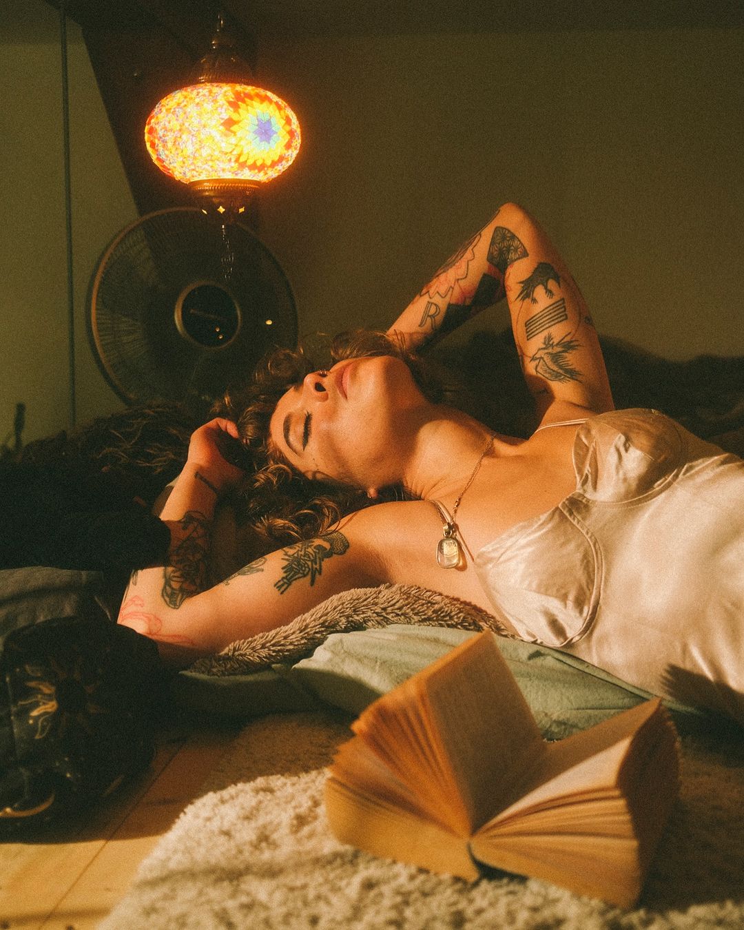 Woman with tattoos lounges indoors, illuminated by a colorful hanging lamp. She's surrounded by a book, pillows, and an electric fan.