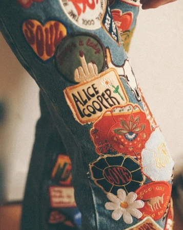 Close-up of blue denim jeans covered in various colorful embroidered patches and appliques, showing a whimsical and artistic design.