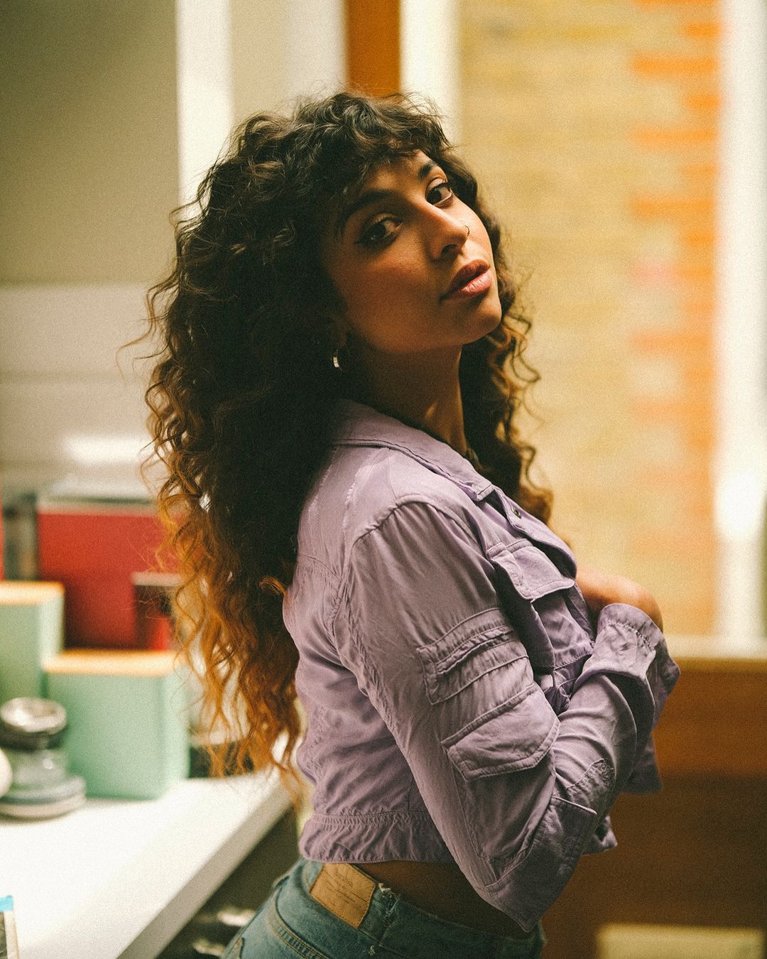 A young woman with curly brunette hair and a light jacket poses indoors in a medium shot with a neutral facial expression.