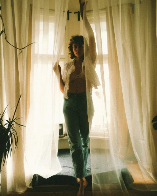 Woman stands barefoot in jeans and lacy top behind translucent white curtains in soft light indoors, with a leafy plant beside her.