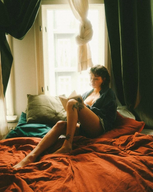 Young woman reading a book in bed by the window. She is barefoot, wearing a shirt and shorts, and has tattoos on her legs.