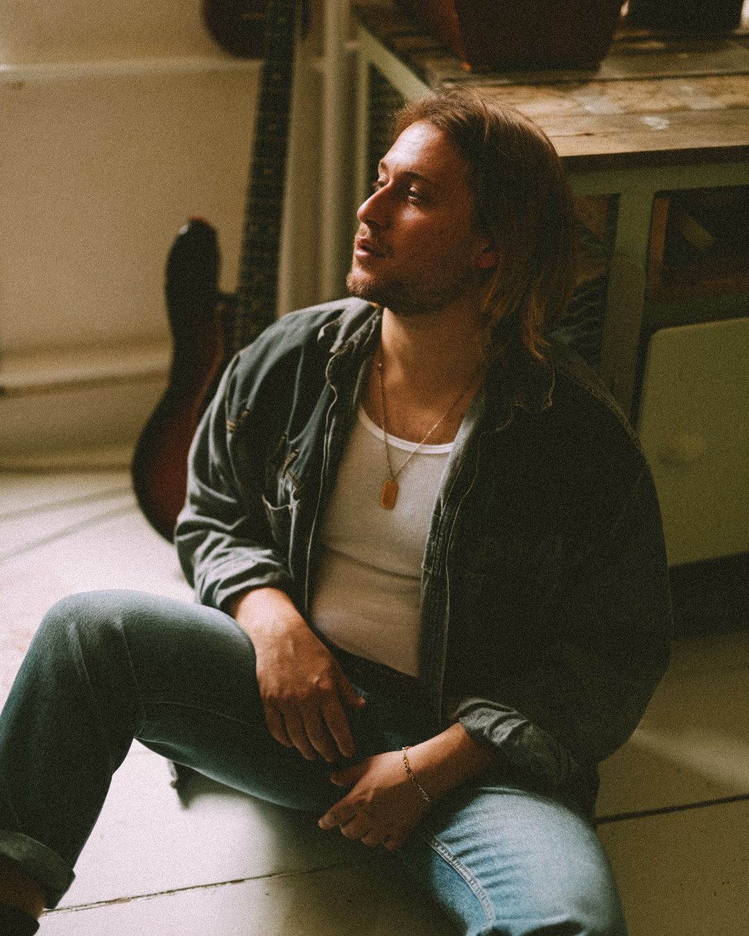 Man sits indoors in casual denim and white t-shirt. Natural window light and nearby guitar. Relaxed pose, looking slightly off-camera.