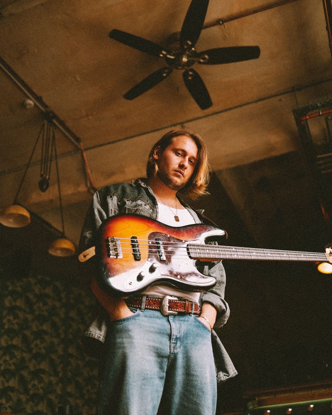 Young man holding a vintage electric guitar in a dimly lit room with a ceiling fan. Wearing a denim jacket and jeans. 