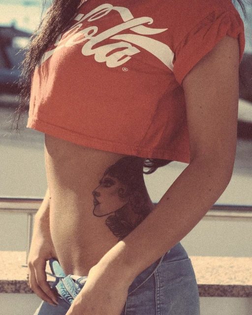 Torso of a woman wearing a cropped Coca-Cola t-shirt and jeans, showcasing a tattoo on her side against an outdoor background.