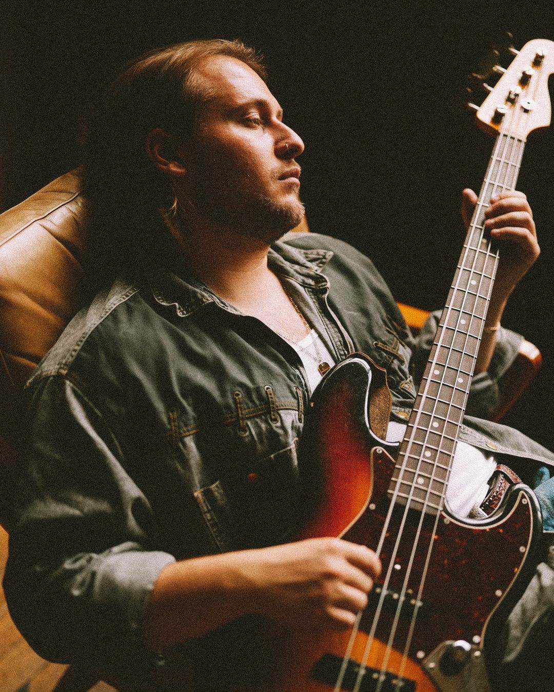 Adult male in casual clothing seated holding an electric bass guitar indoors with dark lighting in a music performance or rehearsal setting.