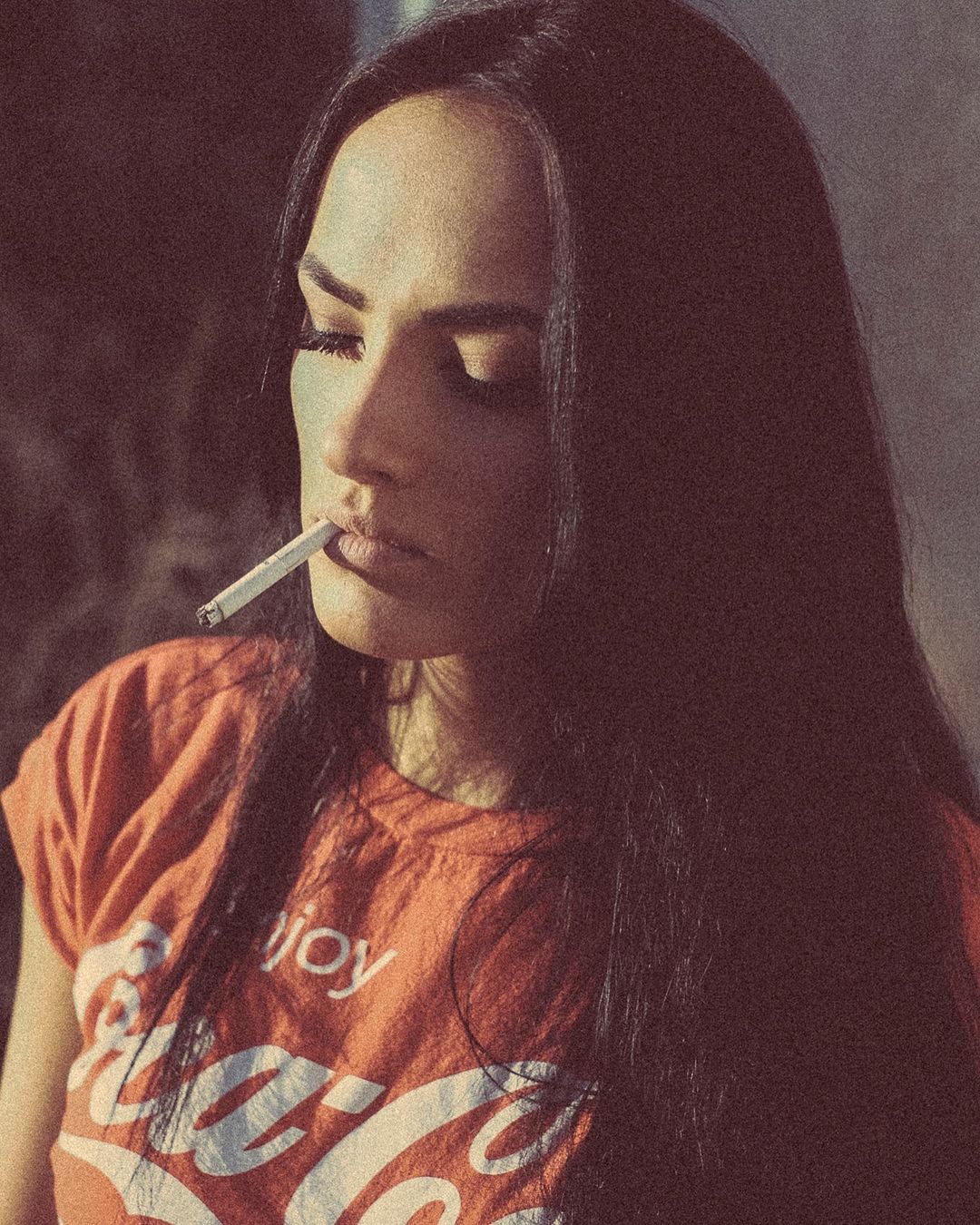 Woman with long dark hair smoking a cigarette. She is wearing an orange Coca-Cola t-shirt, medium shot, warm lighting.