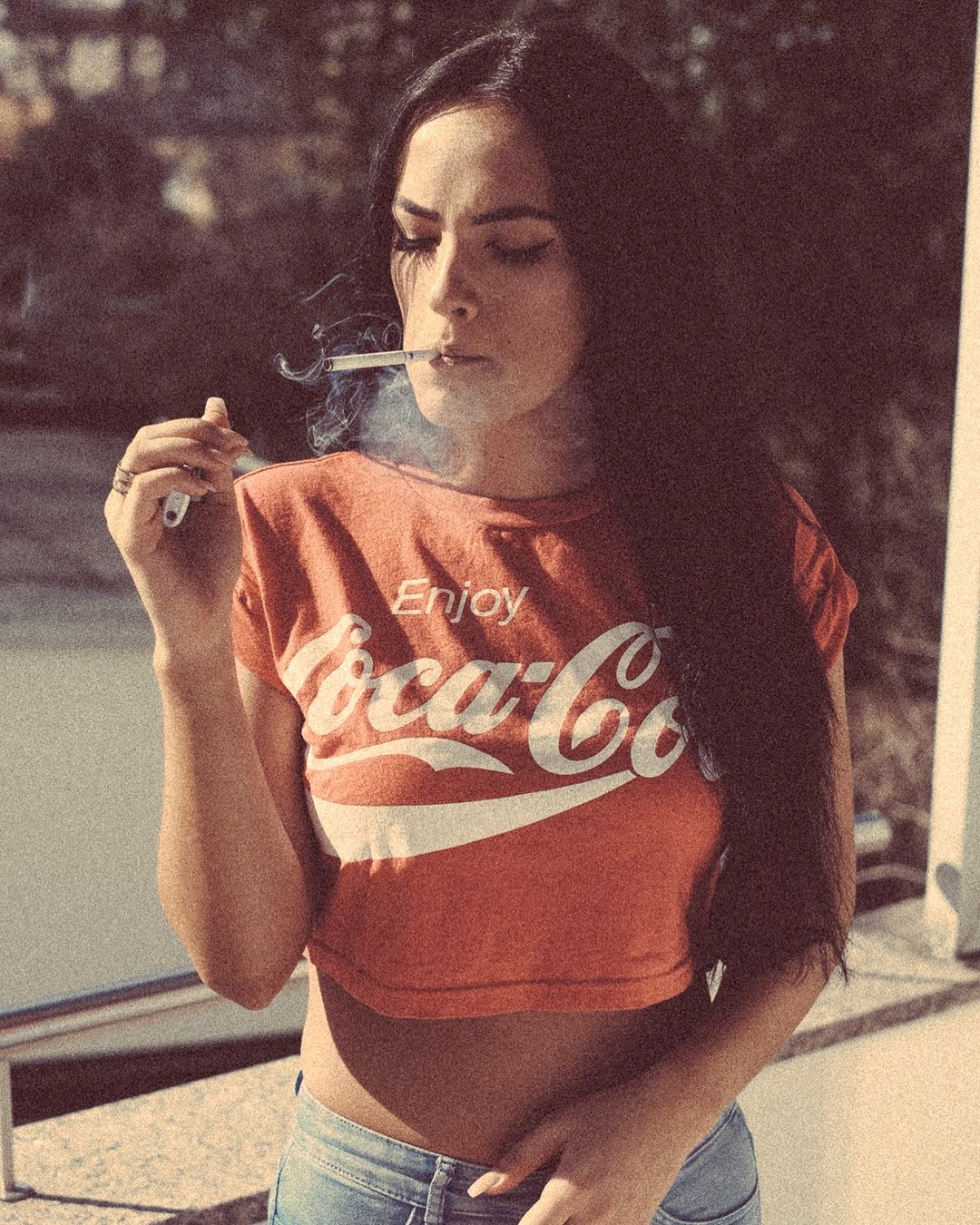 Brunette woman with a Coca-Cola t-shirt, smoking a cigarette outdoors in natural light, with a filtered vintage effect.