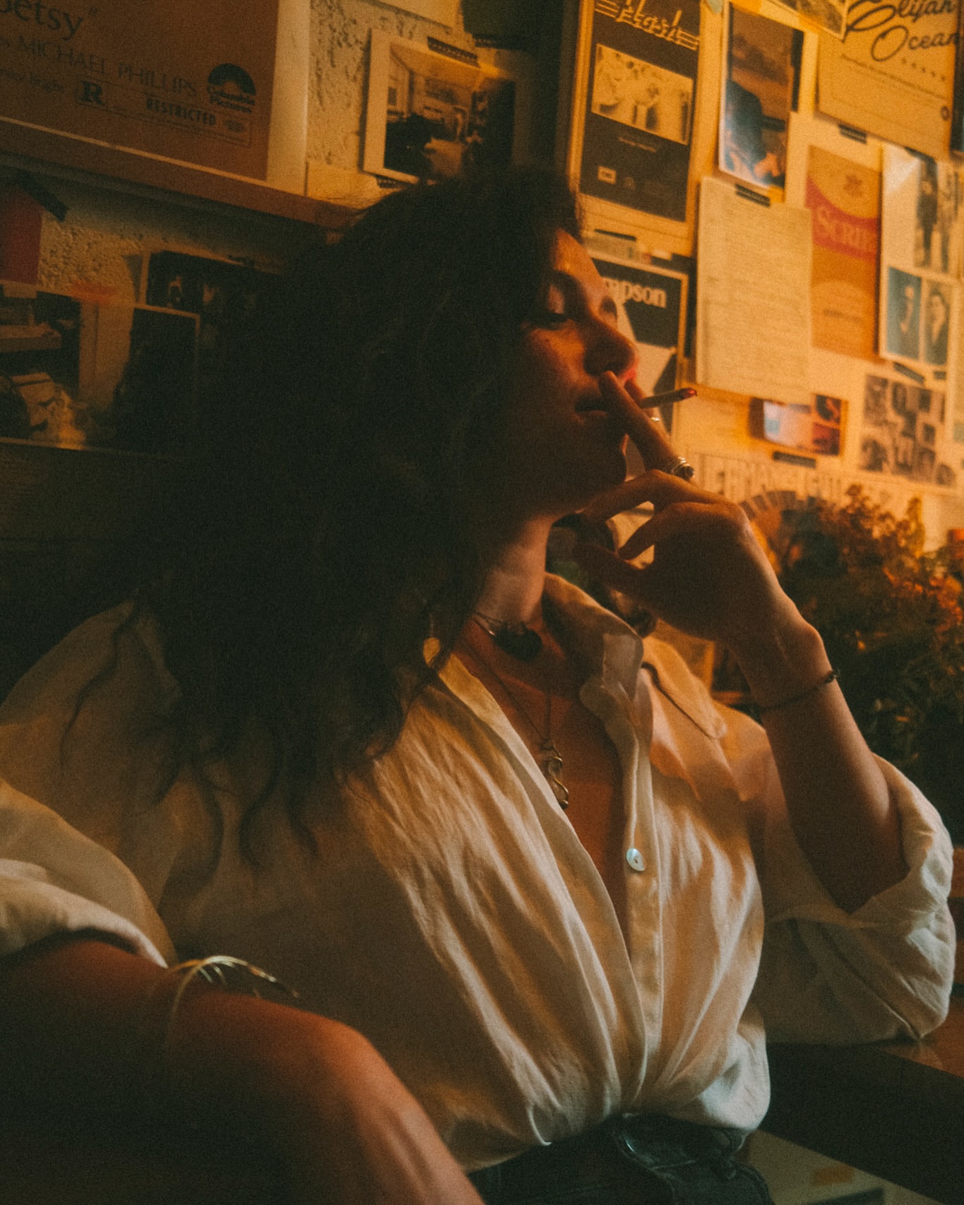 Woman in a white button-down shirt smoking a cigarette against a wall covered in posters