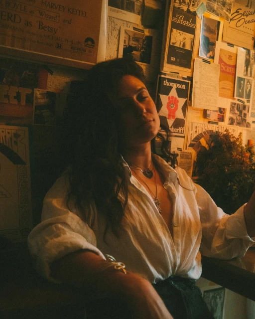 Brunette woman in a white shirt relaxes in a chair against a wall covered in vintage posters and signs in a warm-toned indoor setting.