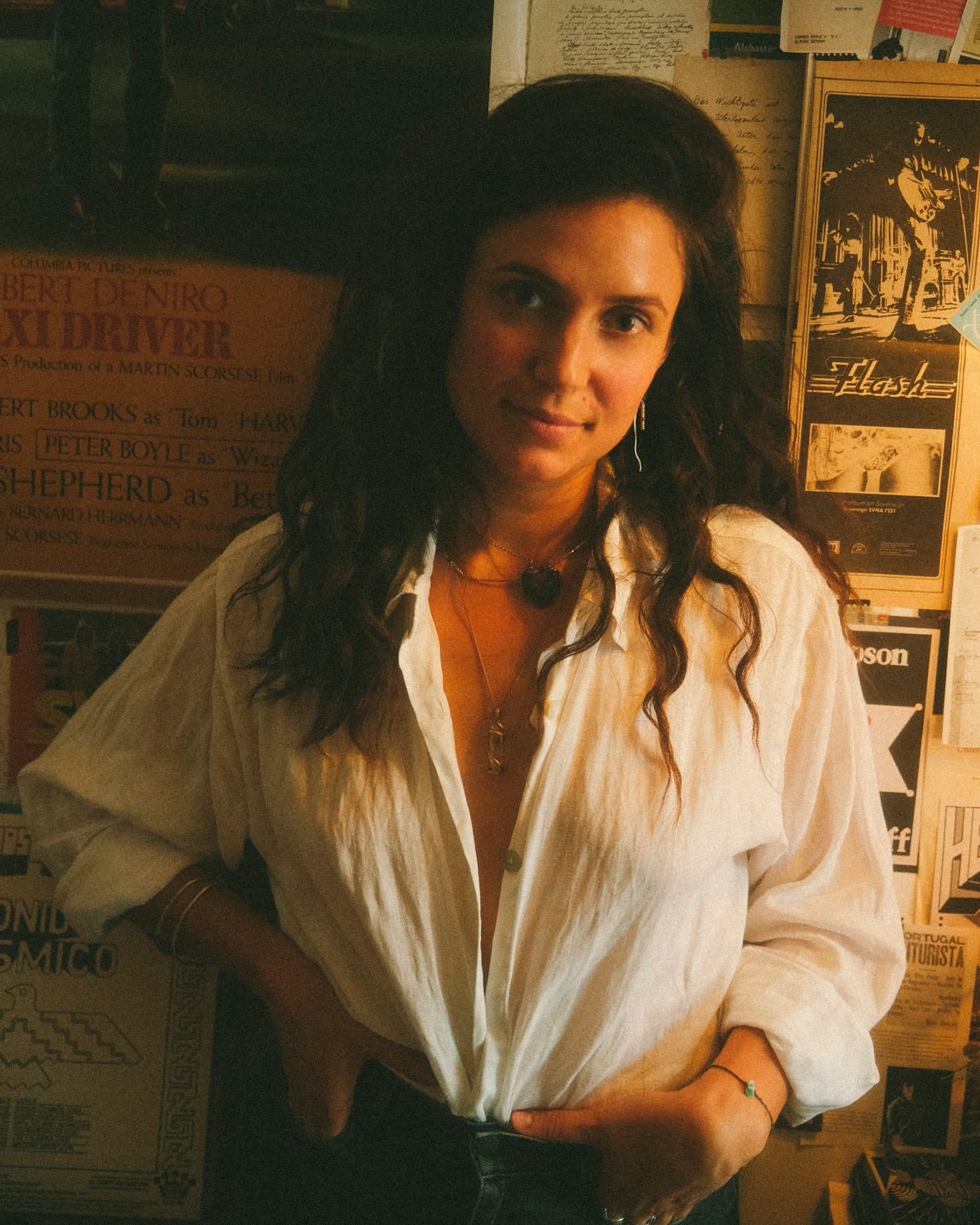 Portrait of a woman in a white shirt with movie posters in the background, posing indoors with her hand on her hip.