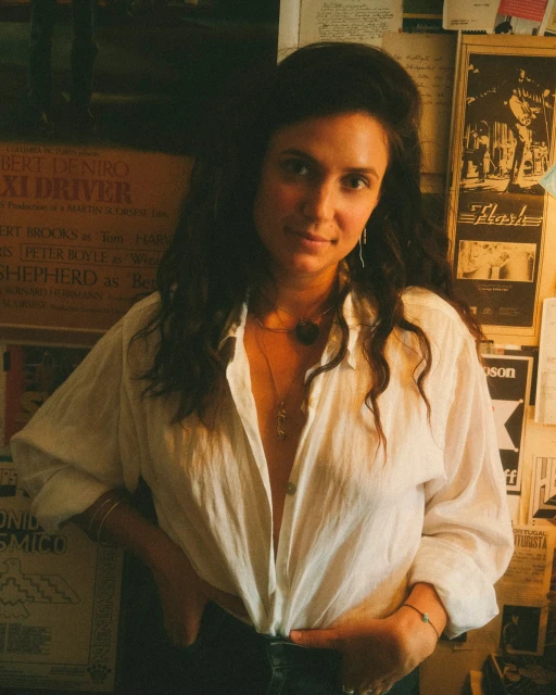 Portrait of a woman in a white shirt with movie posters in the background, posing indoors with her hand on her hip.