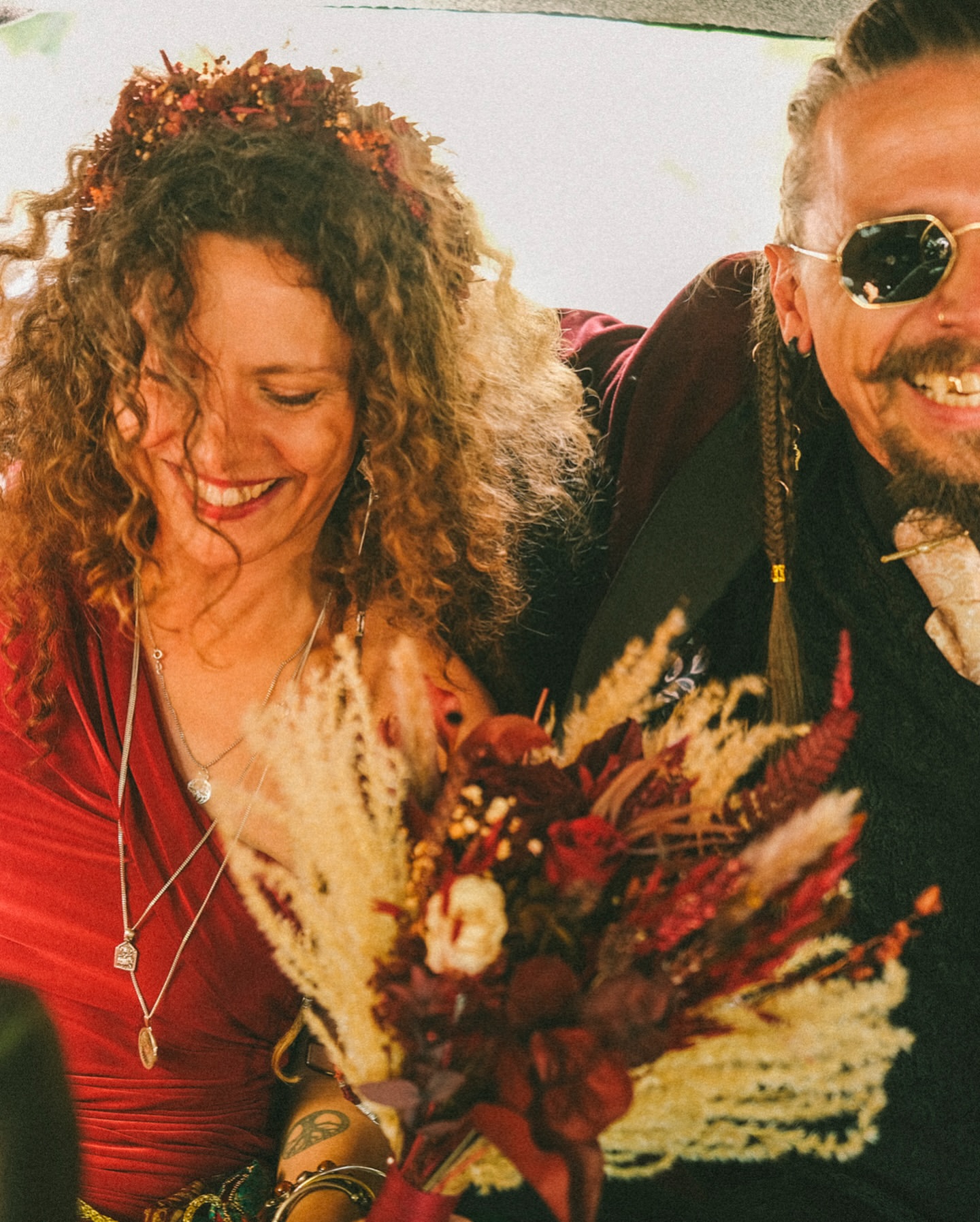 Smiling couple in wedding attire, the woman with a flower crown, holding a bouquet, the man with a mustache and sunglasses.