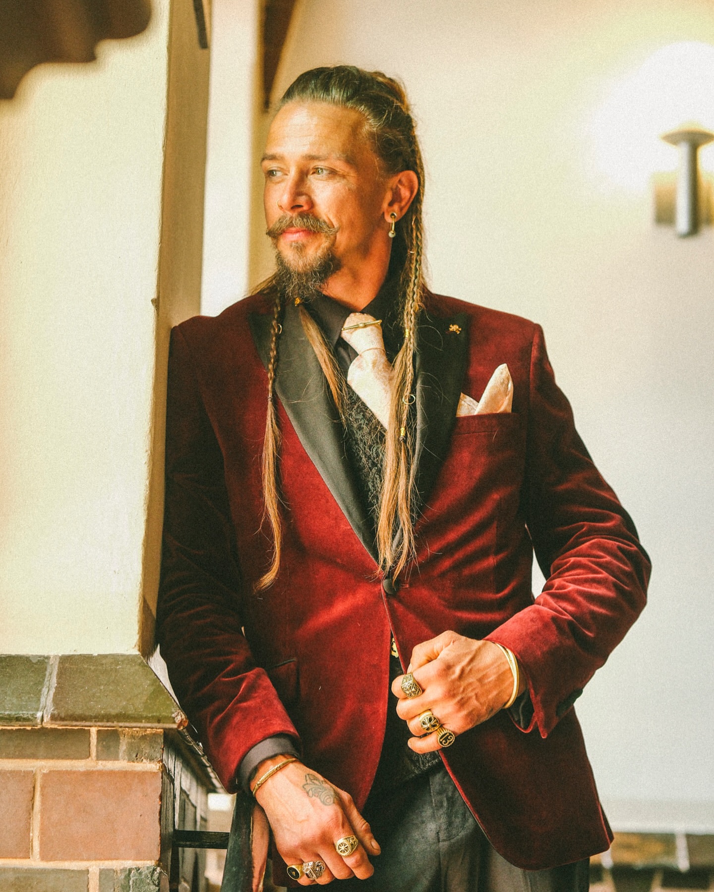 Man in a burgundy velvet blazer poses indoors with intricate braids and rings, creating a fashionable and timeless portrait.