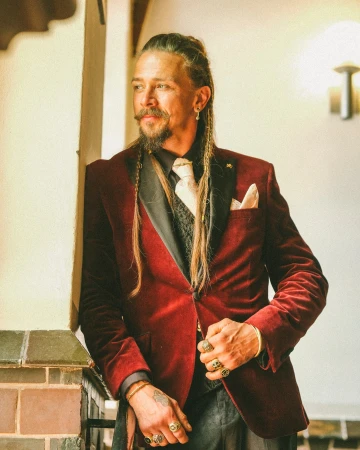 Man in a burgundy velvet blazer poses indoors with intricate braids and rings, creating a fashionable and timeless portrait.
