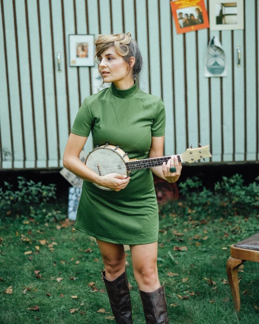 A young woman in a green dress standing outdoors, playing a banjo, with a vintage trailer and grass in the background.