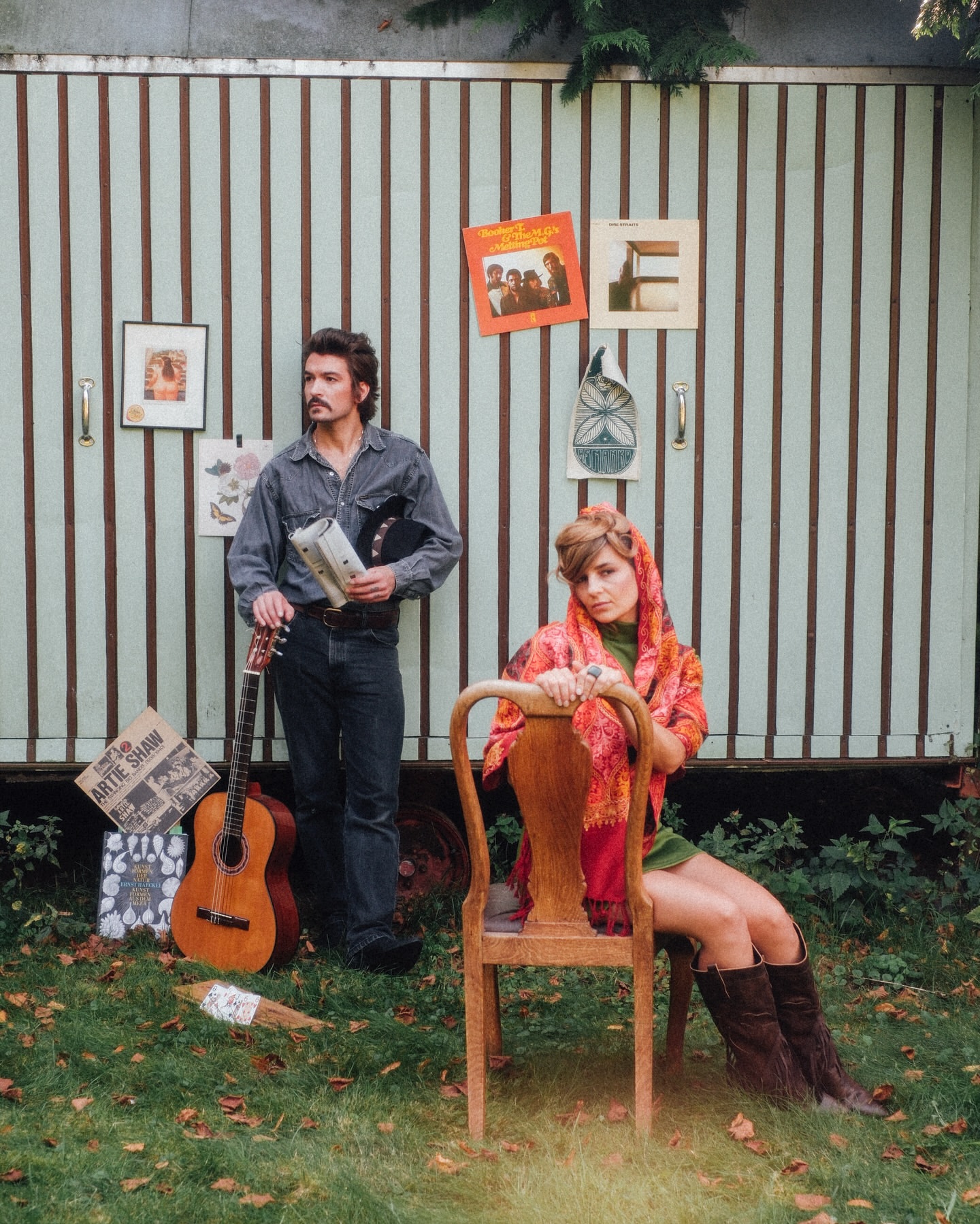 Retro-styled photo of a man with a guitar and a woman seated on a chair, set against a striped wall adorned with vintage records.