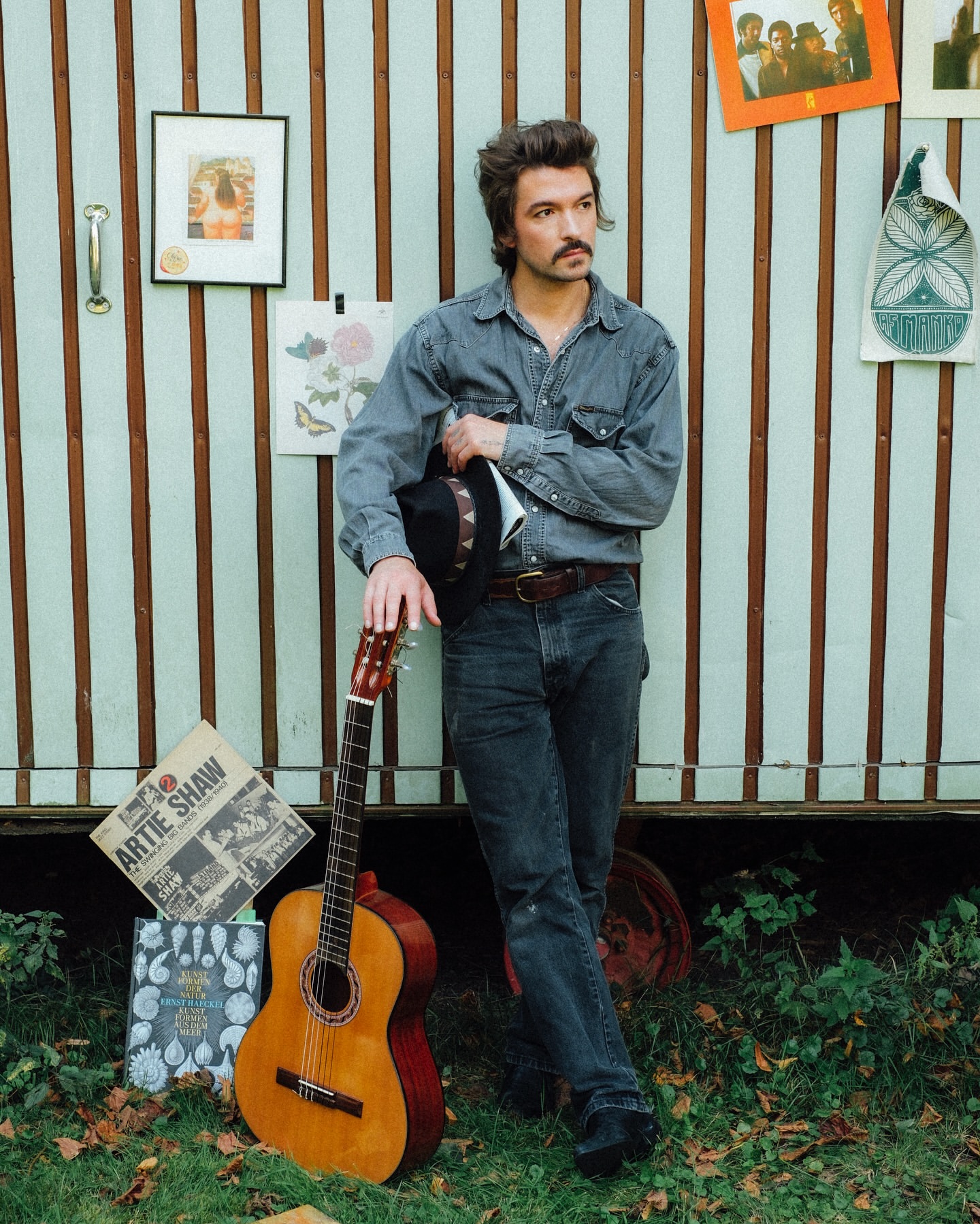 Man in denim shirt holds hat, leans against wall with guitar on grass. Album covers & drawings decorate the background.