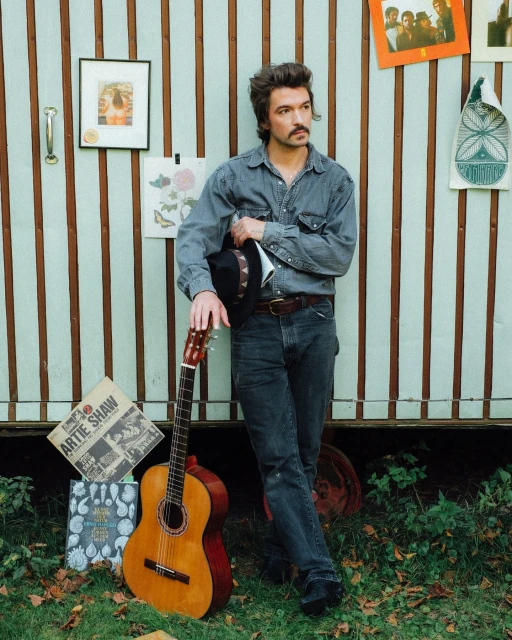 Man in denim shirt holds hat, leans against wall with guitar on grass. Album covers & drawings decorate the background.