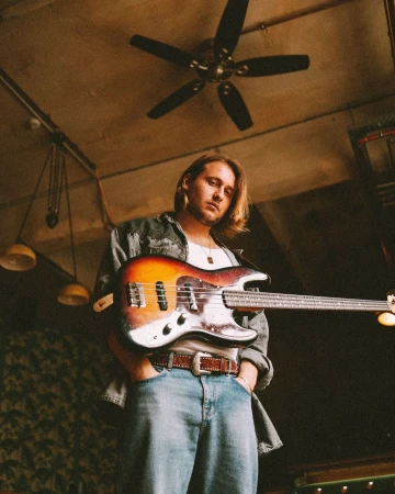 A man with shoulder-length blond hair holding an electric bass guitar in an indoor setting photographed by Bob Sala.
