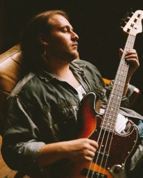 A man in a green shirt holding and playing a bass guitar while seated in a leather armchair.