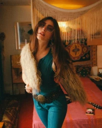 Young woman in a blue jumpsuit with a fur stole and tinted glasses posing in a bohemian bedroom, photographed by Bob Sala.